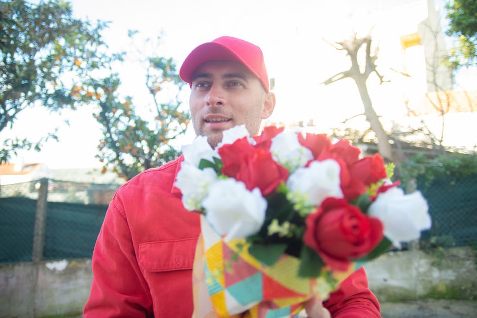 A man wearing a pink cap and red jacket is holding a floral gift consisting of a bouquet with red and white roses, wrapped in colorful paper with a yellow ribbon. He is outdoors, with blurred greenery