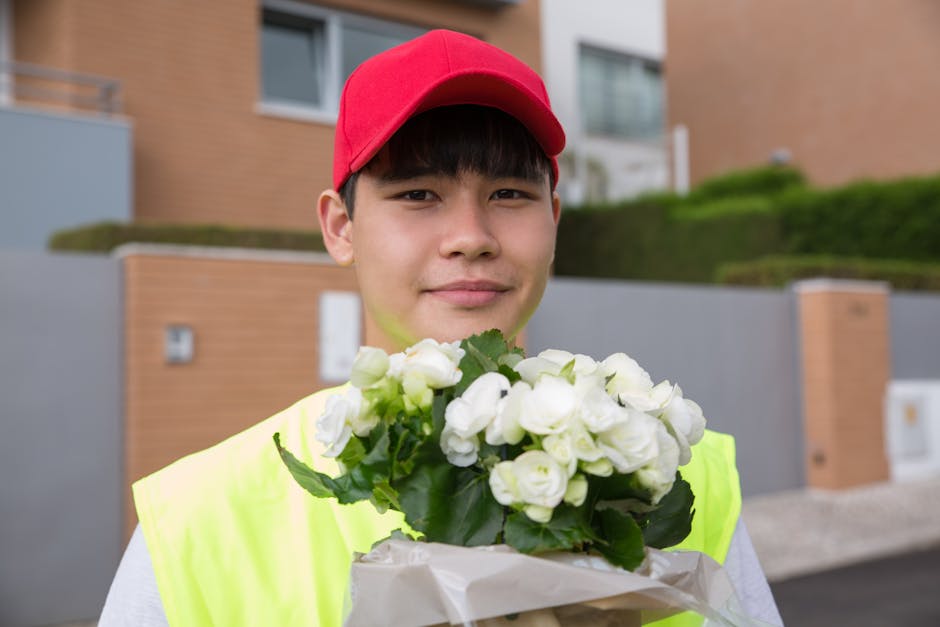 A young male delivery person wearing a bright yellow high-visibility vest and a red cap holds a bouquet of fresh white flowers wrapped in white paper. The flowers include white roses and buds with gre