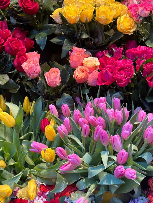 A vibrant display of fresh flowers including pink tulips with green stems and leaves, yellow tulips with similar foliage, and an assortment of roses in shades of pink, peach, and yellow, arranged clos