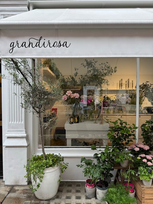 View of a floral boutique named 'grandiroza' with a large white awning above the window, showcasing an elegant and inviting display. Outside, in front of the glass storefront, a variety of potted plan