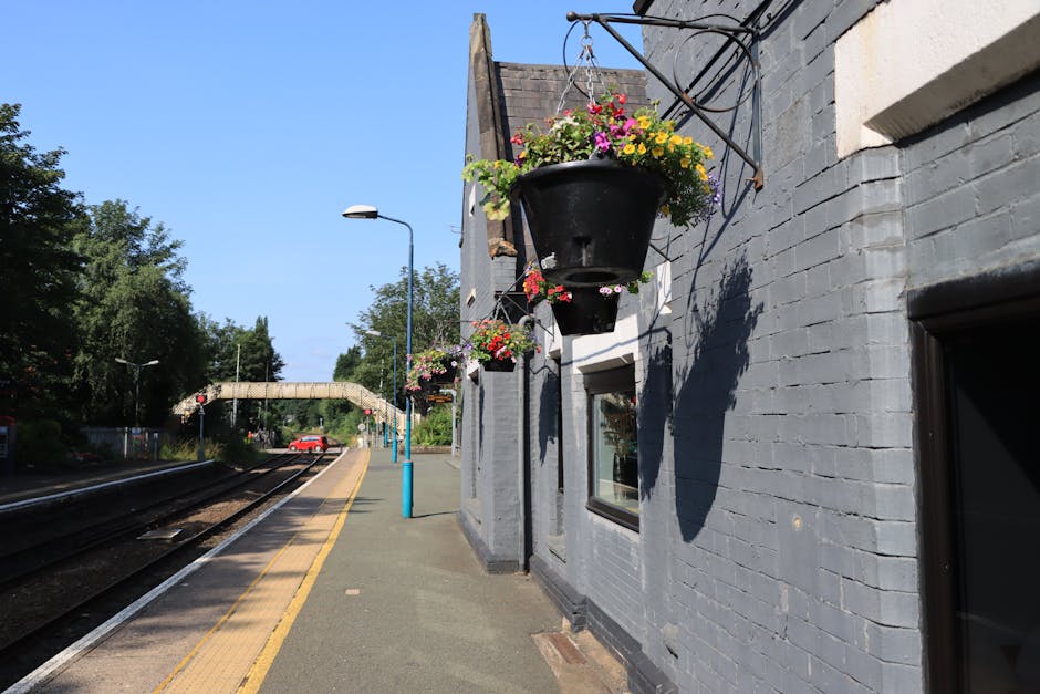 A close-up view of hanging flower baskets attached to the grey brick exterior of a building at Woodford station, featuring vibrant pink, yellow, and purple blooms with lush green foliage. The baskets 