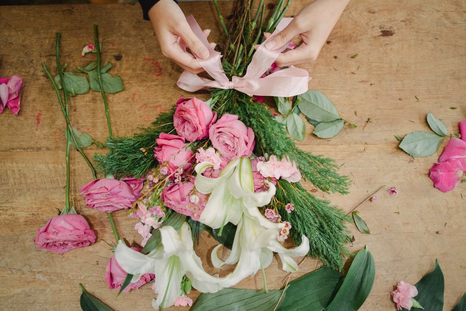 A florist's hands are seen arranging a floral bouquet on a wooden table. The bouquet features a mix of pink roses, white lilies, and greenery including sprigs of conifer and eucalyptus leaves. Some ro