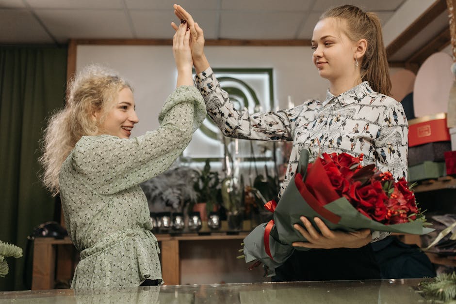 Two women inside a floral shop engage in a celebratory gesture, with one woman holding a bouquet of deep red roses wrapped in green paper and tied with a red ribbon. The other woman, smiling, reaches 