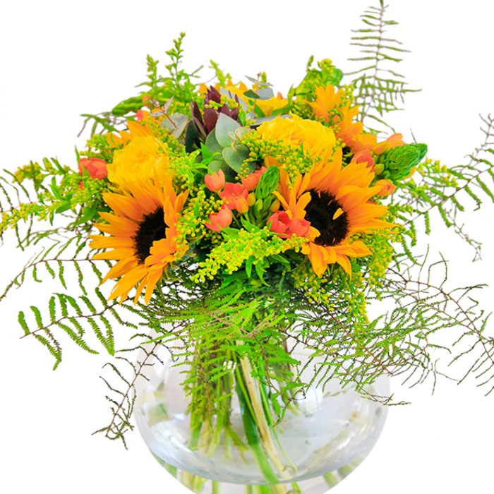 Sunshine and happiness seem to pour from this lavish bouquet, a tribute to Woodford's lively gardens and summer festivals. Vivid yellow sunflowers and roses bloom side by side, each petal layer unfolding in a generous embrace that radiates warmth throughout the design. Deep green foliage, interlaced with trailing ferns and airy sprigs, reaches outward in all directions, channeling the lush abundance of Chestnut Avenue's community gardens. Dots of cherry-red hypericum berries glint among the greenery, their glossy skins popping brilliantly against the matte florals and cool blue-green eucalyptus leaves. A gently curved glass vase, simple yet elegant, cradles every stem, spotlighting the natural architecture and exuberance above. Up close, textures come alive: sunflowers with their suede-like faces, roses furled tightly then blooming wide, and crisp, just-clipped greens. The whole arrangement carries a light, invigorating fragrance-subtle rose and bright foliage-recalling an open window on a breezy morning in Woodford. This bouquet, thoughtfully composed by local hands, is perfect for birthdays, anniversaries, and moments where joy and optimism should take center stage, filling homes from South Woodford to High Road with colour and delight.