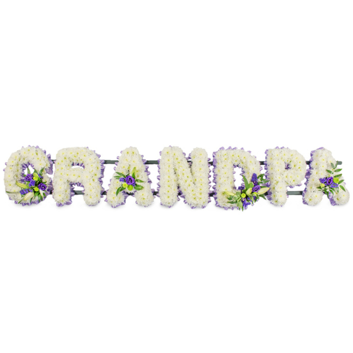 This photograph shows a lovingly crafted funeral flower tribute spelling out "GRANDPA" in large, freestanding letters, aligned in a straight row as though ready to be placed along the front of a coffin or on stands at a Woodford funeral service. Each letter has been built from a sturdy backing, then completely covered with a dense layer of small white blooms-likely chrysanthemums or carnations-that overlap one another to form a plush, continuous texture. The flowers are pure white with a faint creamy warmth at their centers, catching the soft studio light and giving the tribute a peaceful glow, like fresh snow under a quiet winter sky. To break the expanse of white and add depth, the florist has woven in tasteful accents of rich purple and lavender flowers, accompanied by small tufts of fresh green foliage. These coloured clusters are thoughtfully placed: at the lower curve of the "G", within the triangular hollows of the "A"s, and on the upright strokes of the "D" and "P", drawing the eye gently along the name without overpowering it. A deep green metal or foam bar runs just beneath the row, subtly visible in places, binding the letters together and ensuring they stay aligned when moved to churches and crematoriums around Woodford, including those close to George Lane or the High Road. The design exudes a sense of love, honour, and gratitude, a floral echo of memories of a grandfather who might have walked local streets, shared stories over tea, or watched grandchildren grow. It offers families a personalised way to say goodbye, combining classic funeral flowers with a message spelled out in blooms.