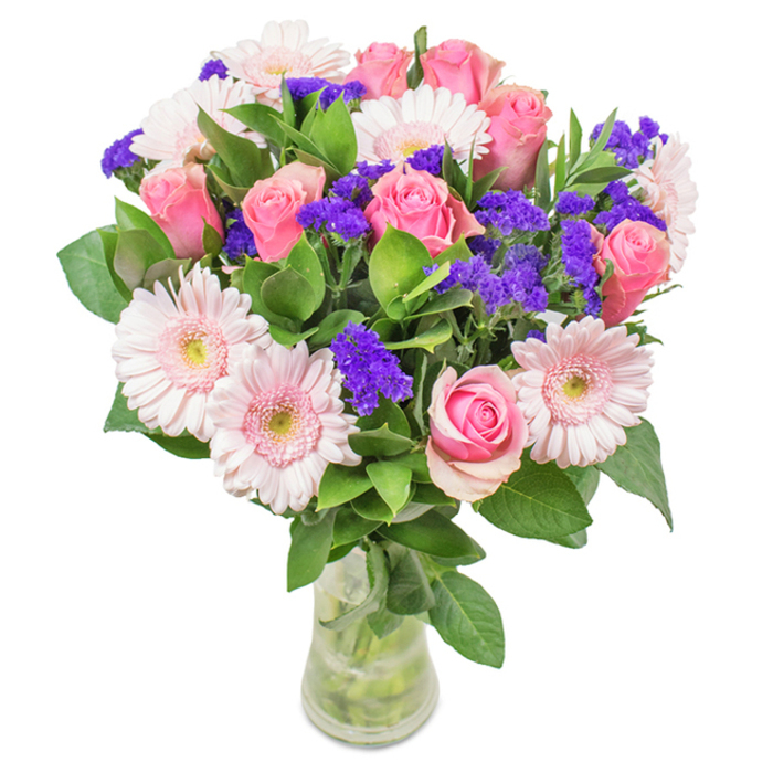 Presented in a crystal-clear glass vase against a pristine backdrop, this vibrant floral arrangement sings with the spirit of Woodford's gardens. At its heart, approximately 7-9 soft to medium pink roses gather, a few still tightly furled while others open in classic spiral elegance-every one chosen for its unique freshness and poise. Interlaced among them, 5-7 delicate pale pink gerbera daisies radiate from round heads, their nearly white centers glowing with quiet charm. Around these, clusters of vivid purple statice bring vivid texture and a burst of colour, reminiscent of the varied blooms found in local Woodford Green flower markets. Deep green foliage wraps the base in a lush embrace, accentuating the arrangement's vibrancy and lending the natural scent of an early-morning walk through Ashton Playing Fields. The stems, visible in clear water within the vase, reinforce a sense of transparent simplicity and show the bouquet has just been arranged for delivery. Whether marking birthdays, anniversaries, or Mother's Day for families throughout Woodford, or serving as a daily accent for a welcoming Oak Hill Road home, this elegant mix of gerberas, pink roses, and purple filler flowers overflows with freshness, affection, and celebration of the local area.